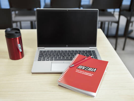 A laptop computer next to a thermos and a notebook with the Riveria logo.